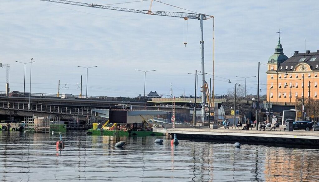 Det pågående bygget av en ny bro för fotgängare och cyklister från Gamla stan till Slussen över Söderström i Stockholm i slutet av april 2026. Foto: Sinikka Halme/Wikimedia Common/https://commons.wikimedia.org/wiki/File:Sderstrm_ny_gng-_och_cykelbro_Nya_Slussen_01_april_2026.jpg