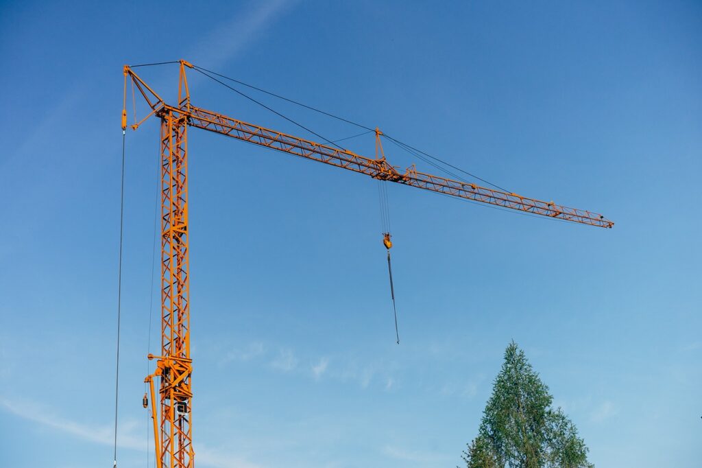 En orange byggkran mot blå himmel med en trädtoppssilhuett i nedre kanten.