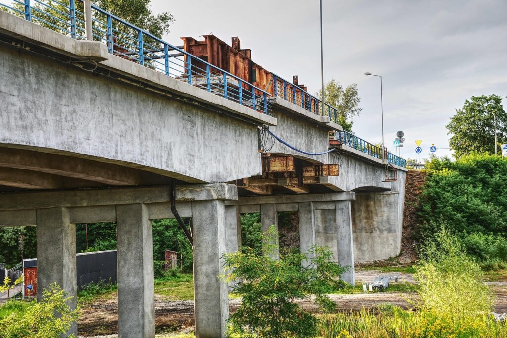 En betongbro med blå räcken och rostiga stålkonstruktioner ovanpå, sedd från sidan med stödben och grön vegetation under bron.