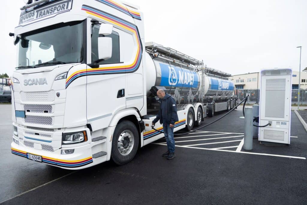 En vit Scania-lastbil med Mapei- och Ruuds Transport-märkning står ansluten till en laddstation medan en person kopplar in laddkabeln.