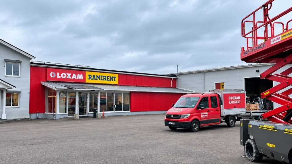 Exteriör av Ramirents och Loxams nya kundcenter i Borlänge med röd fasad, skyltar, en servicebil och en lift på gårdsplanen.