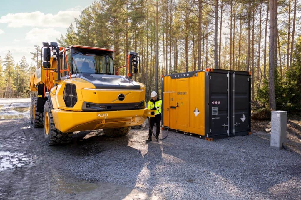 En gul Volvo A30 Electric-dumper står bredvid en laddcontainer i en skogsmiljö medan en person ansluter laddkabeln.