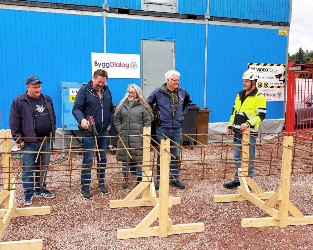 Fyra personer står vid en armeringskonstruktion på en byggarbetsplats framför en blå byggnad med ByggDialog-skyltar.