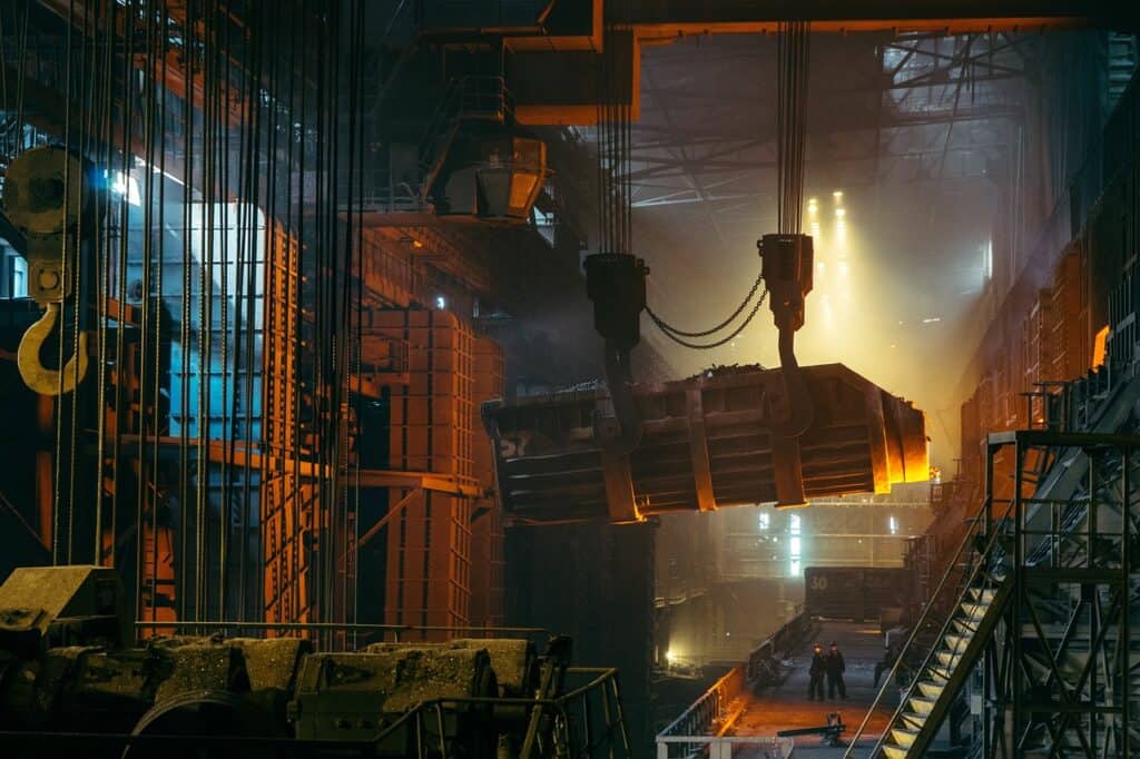 En industrimiljö med en stor kran som lyfter ett glödande stålämne i en fabrikshall.