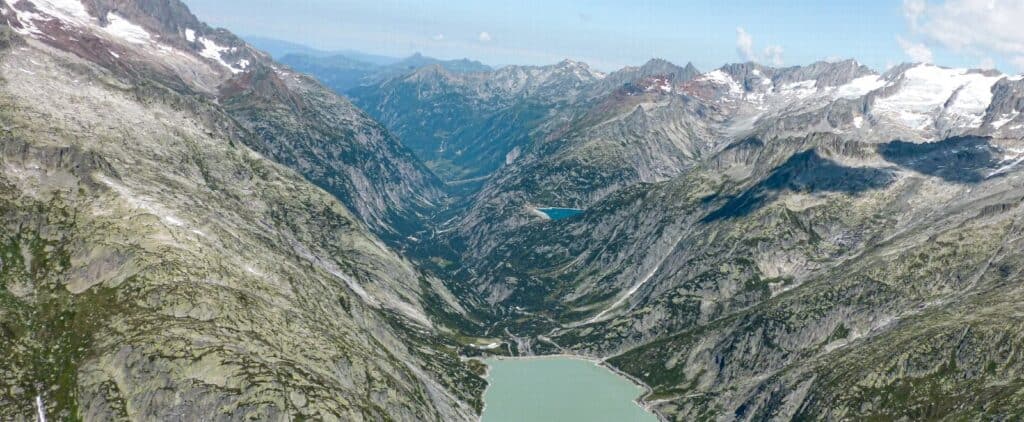 Flygbild över en alpdal med höga berg, en sjö i förgrunden och bergsmassiv på båda sidor.