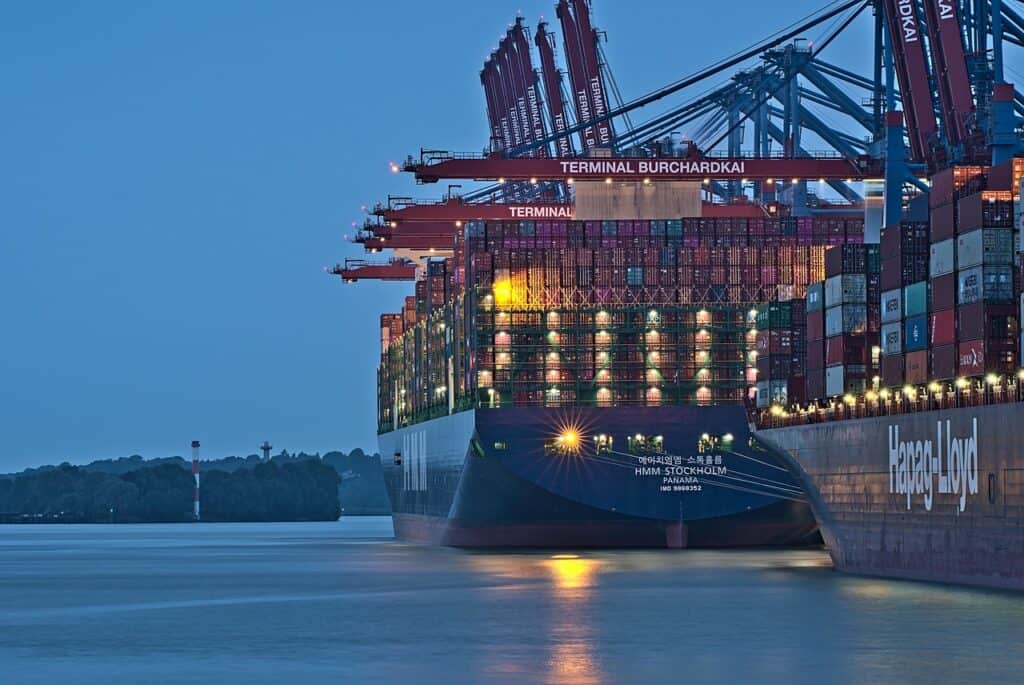 Containerfartyg vid kaj med stora hamnkranar och staplade containrar i bakgrunden.