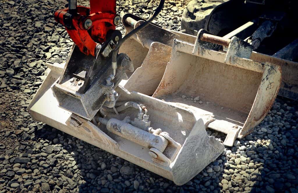 En grävmaskinsskopa med hydraulcylinder och slangar står på grov makadam på en byggarbetsplats.