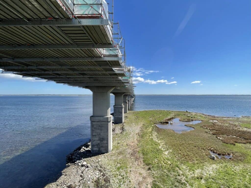 Ölandsbron sedd underifrån med byggställning under brobanan och pelare i havet, med blå himmel och grönt strandområde till höger.