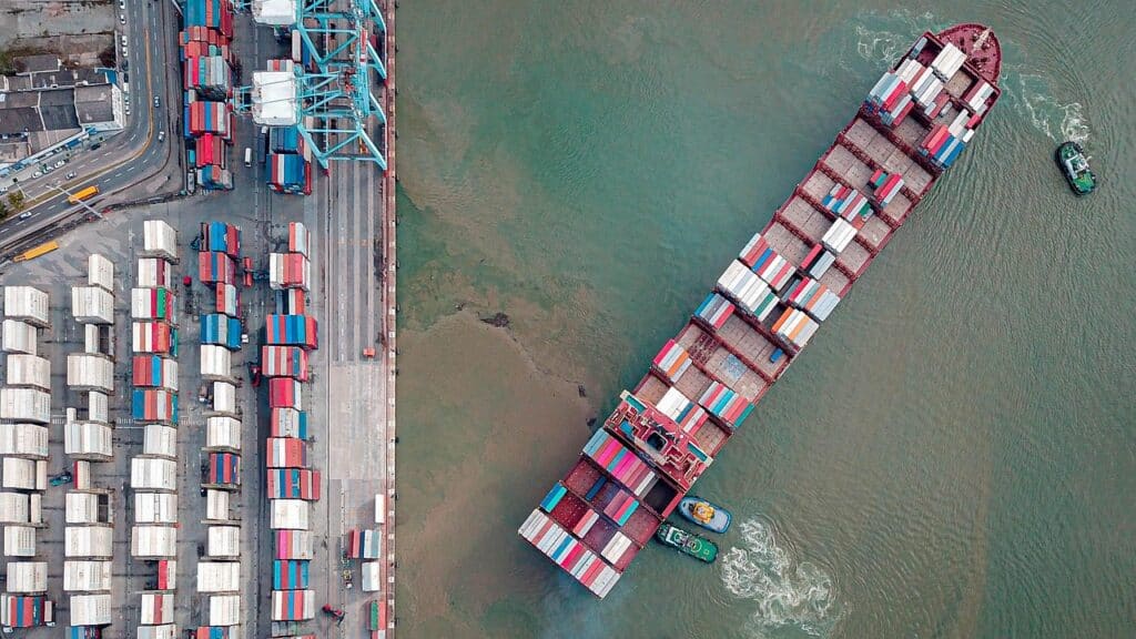 Flygfoto över ett containerfartyg som lämnar en kaj, med bogserbåtar, containerstaplar och kranar i hamnområdet.