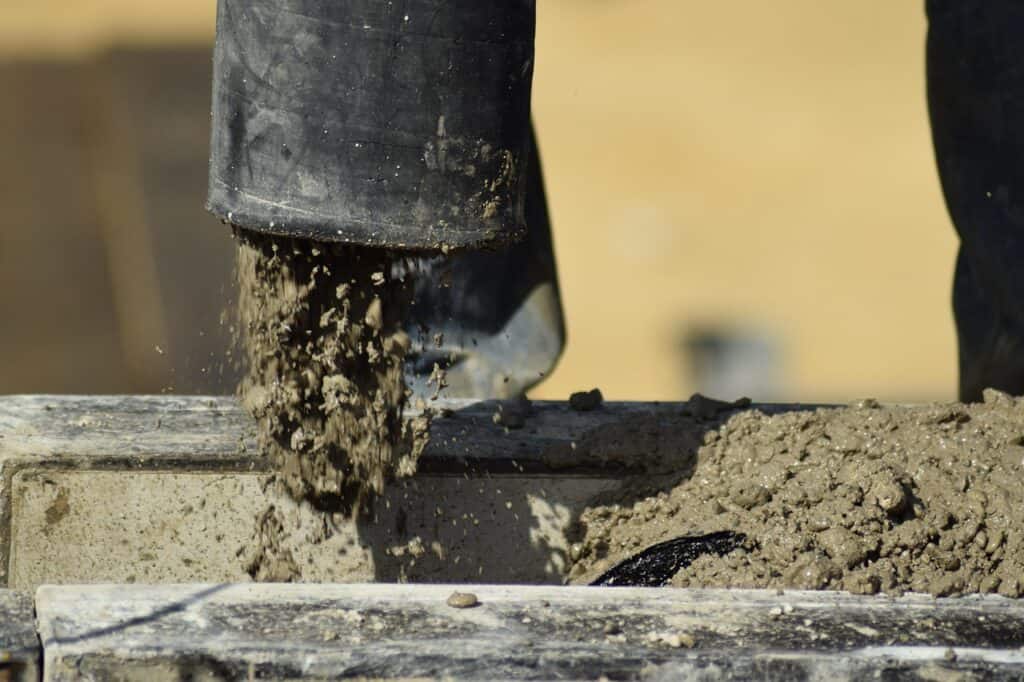 Grå betong som rinner ur en svart gjutslang ned på en betongyta, med sandig bakgrund.