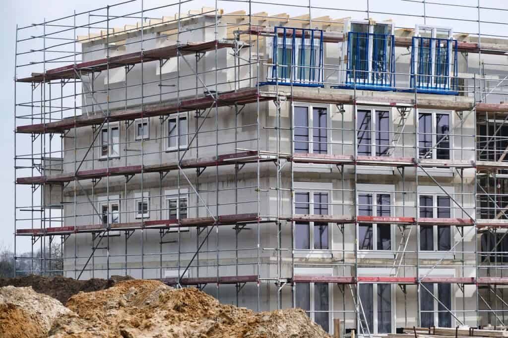 Nybyggt flerbostadshus omgivet av metallställningar, med fönster och grovt schaktmaterial i förgrunden.