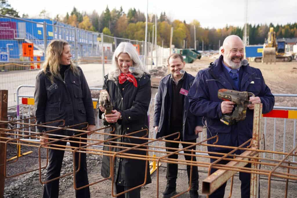Fyra personer står vid en byggplats bakom armeringsstänger av rostigt stål; två kvinnor och två män håller armeringsverktyg, och bakom dem syns ett blått byggstaket och grävmaskiner.