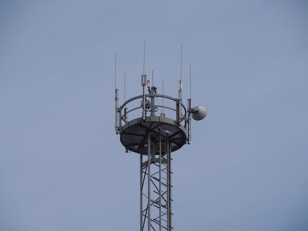 Toppen av ett radiomast med antenner, en parabolantenn och en plattform med räcke mot en grå himmel.