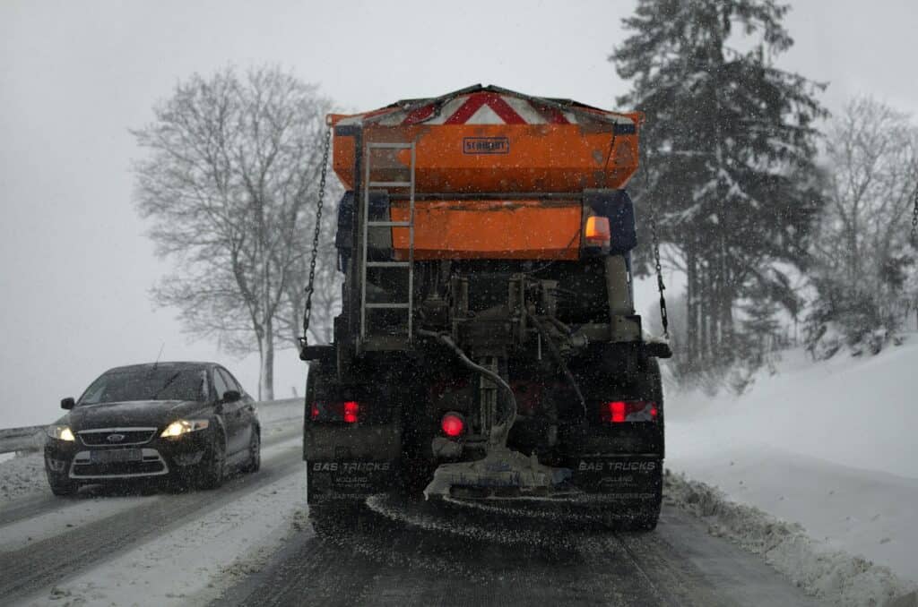 Orange saltspridare på en snöig väg i snöfall, med en bil med tända strålkastare framför.