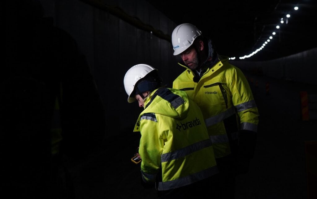 Två personer i gul varselväst och vita skyddshjälmar med Bravida-logotyp står i en mörk tunnel med takbelysning. Den ena tittar ner mot en handhållen enhet.