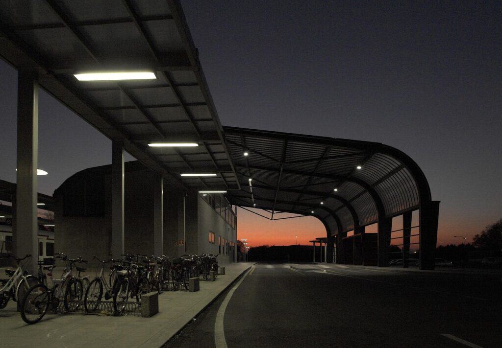 Modern bussterminal med böjd kanopi och belysning i skymningen, cyklar parkerade vid ena sidan och en tom körbana framför.
