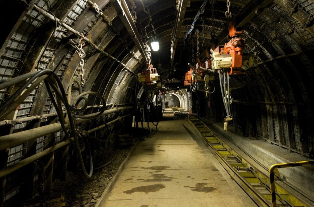 Underjordisk gruvtunnel med rör, kablar, orange hängutrustning och räls under en tunnelvalv.