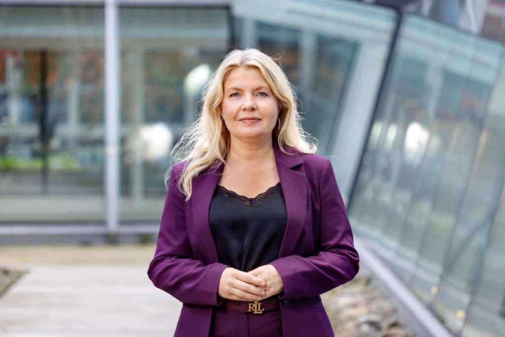 Kvinna med långt blont hår i lila kavaj och svart blus står framför en glasfasad och håller händerna framför sig.