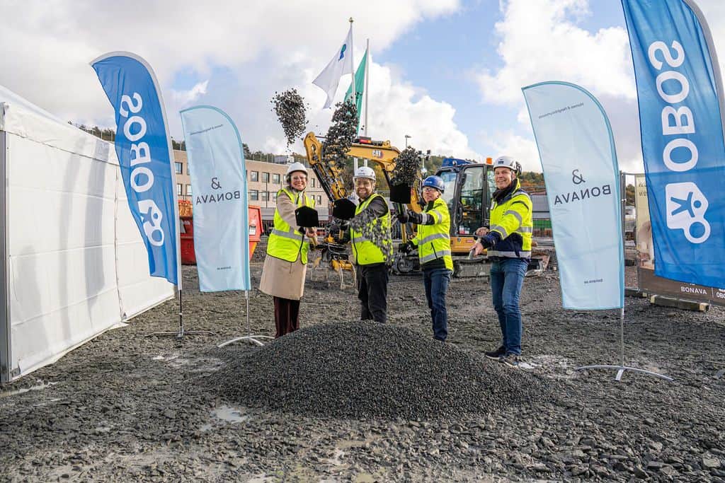 Fyra personer i varselvästar och skyddshjälmar kastar grus med spadar framför en grävmaskin och blå banderoller med texterna OBOS och Bonava.