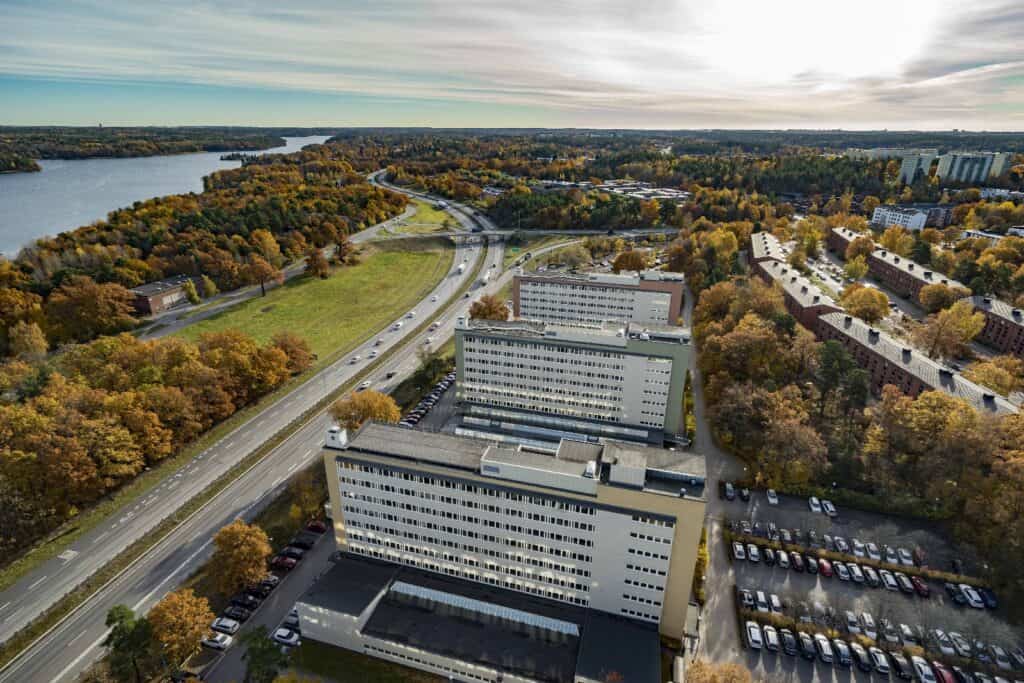 Flygfoto över Telestaden med kontorshus i flera våningar, en väg med bilar, parkeringsplatser, träd i höstfärger och sjö i bakgrunden.
