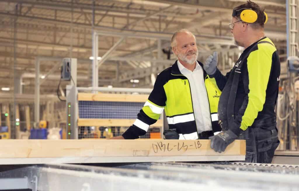 Två män i gul-svarta varselkläder står i en industrihall och pratar. Den ena håller handen mot en träbalk med texten 'OIIC-LS-18'; den andra bär hörselskydd och skyddsglasögon och gestikulerar.