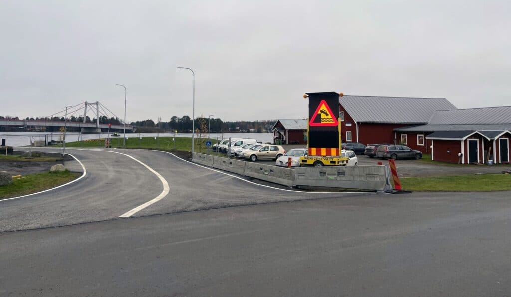 Nybyggt färjeläge i Strömsund med asfalterad väg, betongbarriärer, en varningsskylt för färja och bil på parkeringen, bron i bakgrunden.