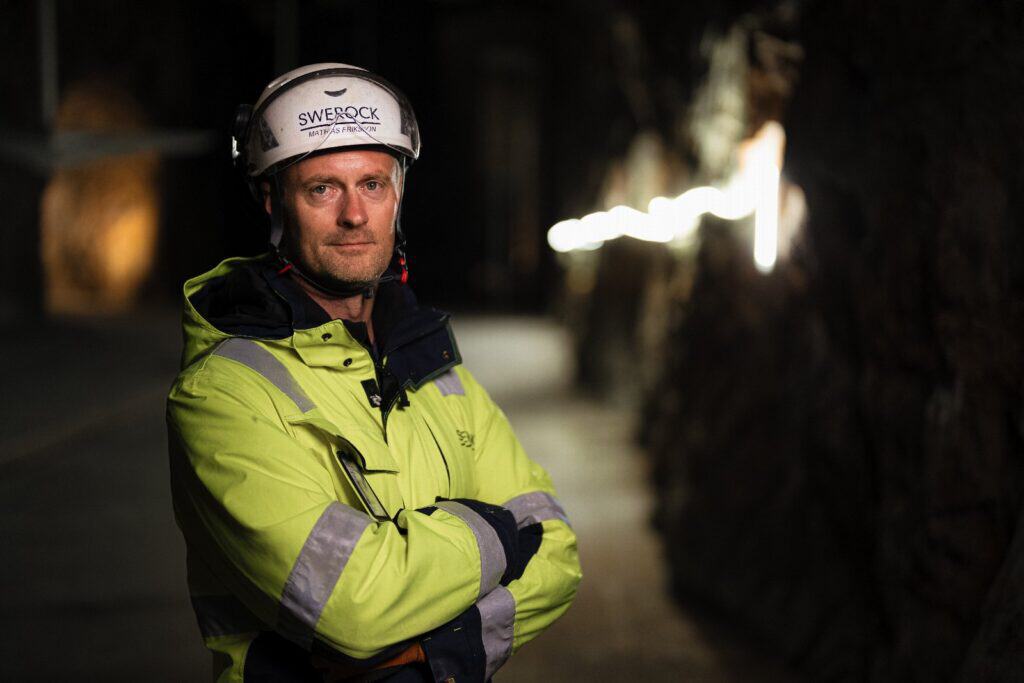 Man i vit hjälm med texten Swerock och Mathias Eriksson och gul varselväst med reflexband korsar armarna i en mörk tunnel.