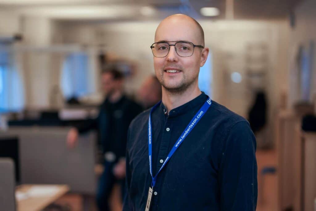 Man med glasögon och mörk skjorta, blått band med texten Lambertsson, står i ett kontorslandskap och ler mot kameran.