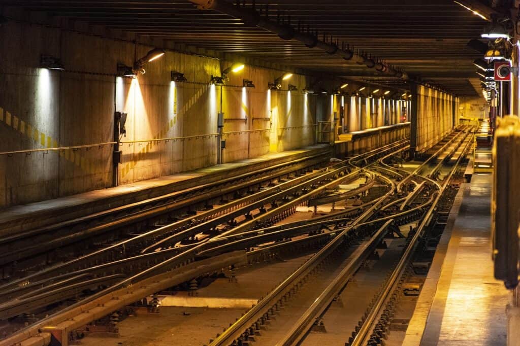 Tunnelbanespår med växlar och skenor i en underjordisk tunnel, upplyst av gula lampor längs väggen.