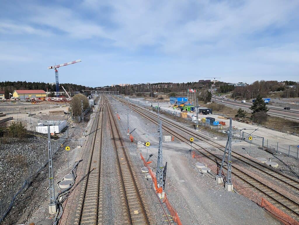 Flera spår med kontaktledningsstolpar och bygge i förgrunden, en kran till vänster och en motorväg till höger i Barkarby.