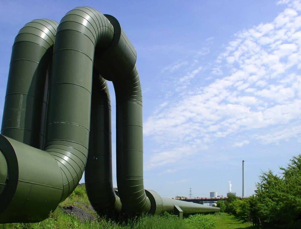 Gröna, böjda industrirör som går över ett gräsbevuxet område mot en blå himmel med vita moln, med ett industriverk och en skorsten i bakgrunden.