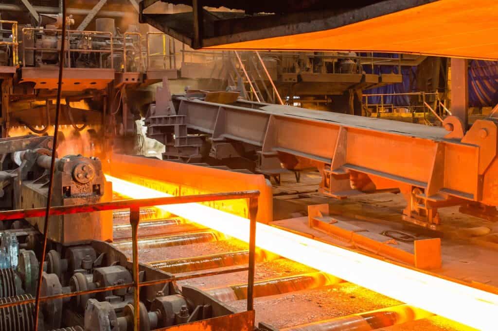Glödande stålbana i ett valsverk med rullar och industriella maskiner i bakgrunden.