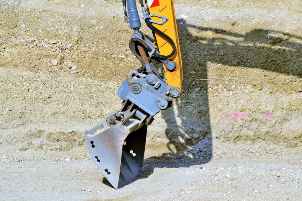 En gul grävmaskinsarm med en grå skopa och vridbar tiltrotator som hänger över ett grusigt byggplatsunderlag.