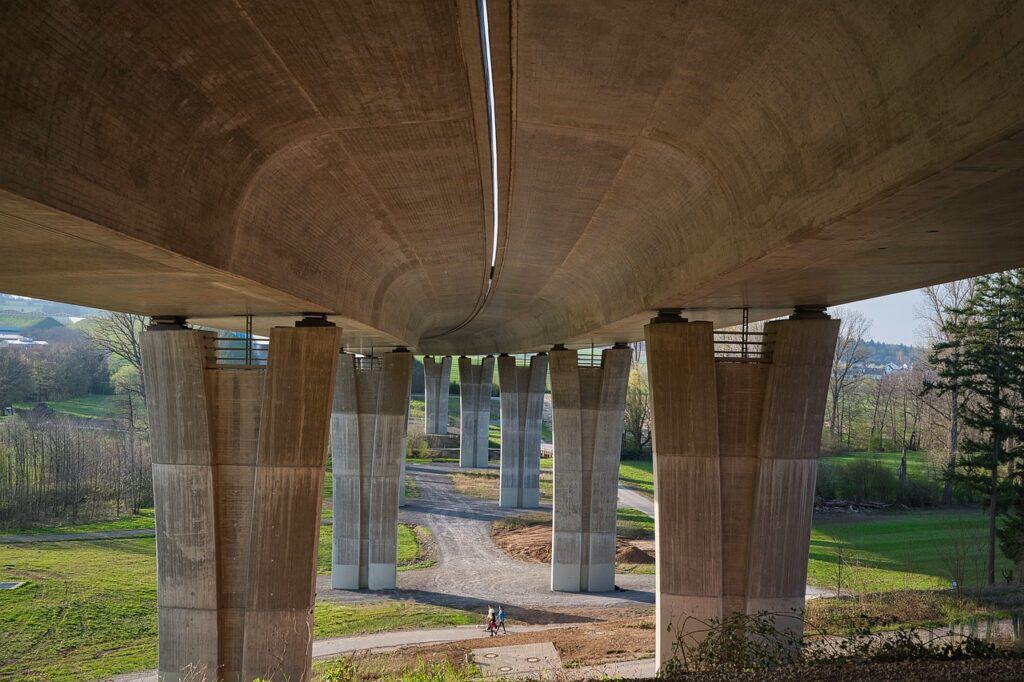 Betongbro sedd underifrån med flera pelare och en gångväg under bron, omgiven av grönska och träd.