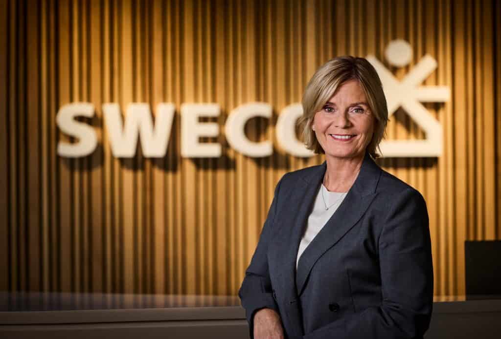 Kvinna med kort blont hår i grå kavaj framför en vägg med vertikala träpaneler och det vita Sweco-logotypen.