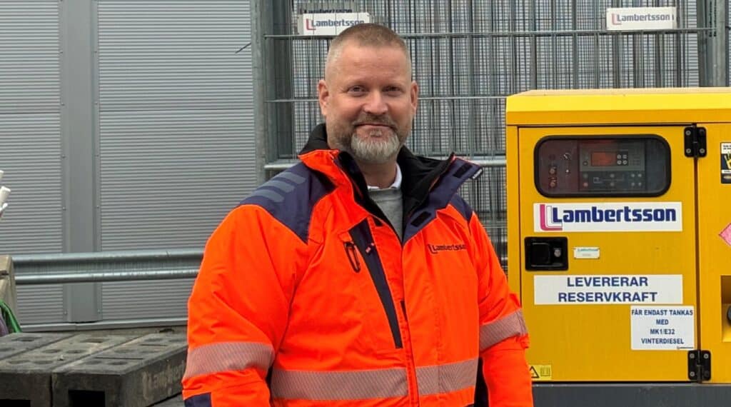 Man med kortklippt hår och grått skägg i orange varsel-jacka med Lambertsson-logotyp, framför ett gult aggregat märkt Lambertsson.