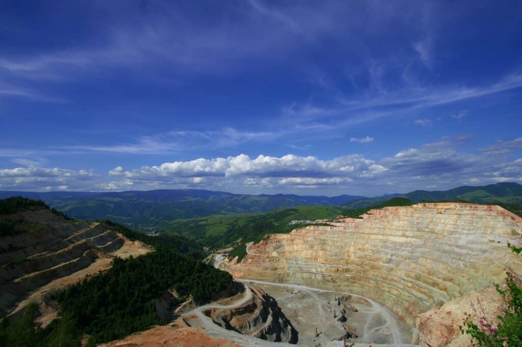 Flygfoto över en öppen gruva med terrasserade bergväggar, slingrande grusvägar och grön skog och bergskedjor i bakgrunden under en blå himmel med moln.
