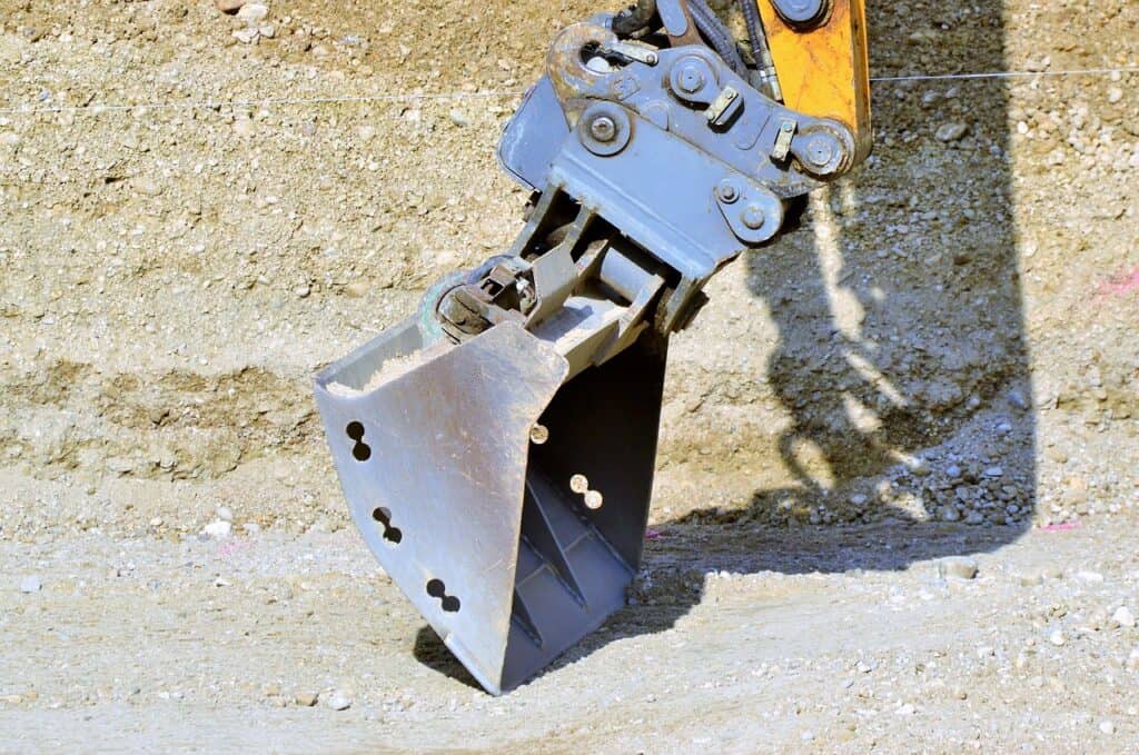 Grävmaskinsskopa med hydraulisk fäste på en byggarbetsplats med grus och sand.
