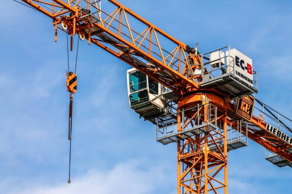Orange tornkran med vit förarhytt och Liebherr-märkning mot blå himmel, med hänglina och lastkrok.
