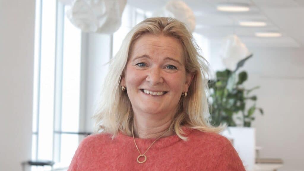 Leende kvinna med blont axellångt hår och rosa tröja samt guldhalsband, i ljus kontorsmiljö med vita lampor och en grön växt i bakgrunden.