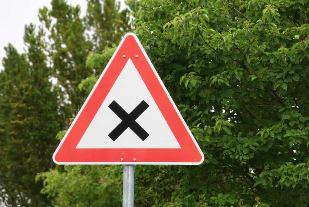 Röd triangulär vägskylt med svart kryss mot grön lövskog och barrträd.
