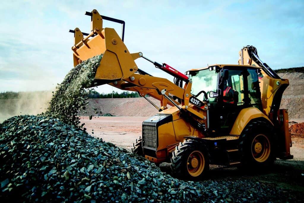 En gul hjullastare från Caterpillar med en skopa full av krossten, som tippas ut på en stor hög av grus och stenar på en byggarbetsplats.